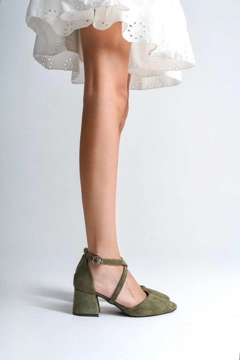 May include: A pair of olive green suede heeled shoes with a criss-cross strap design and a buckle closure.