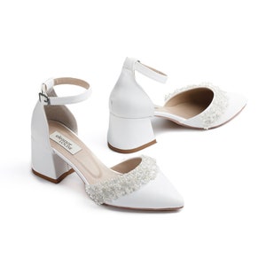 May include: White satin bridal shoes with a beaded embellishment around the toe and a block heel. The shoes have an ankle strap with a buckle closure. The brand name "eleanor rose" is printed on the insole.