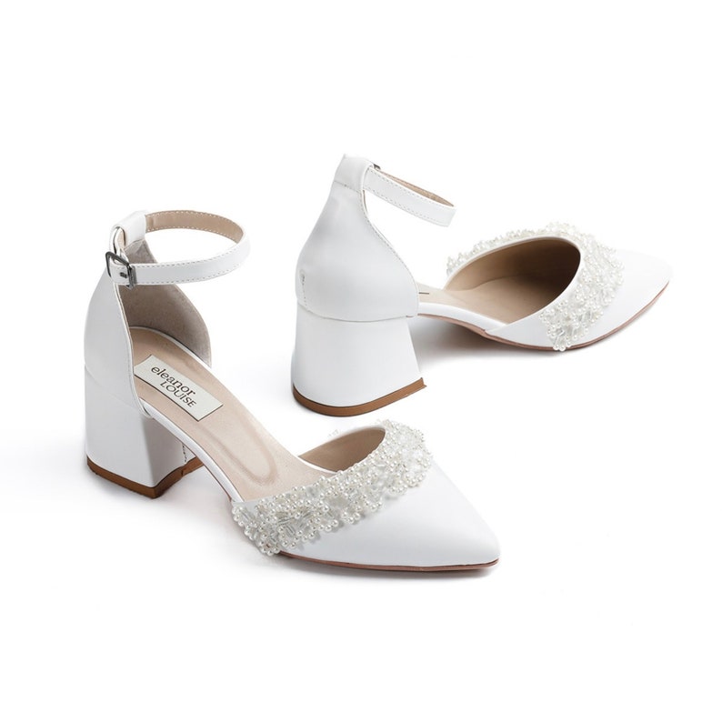May include: White satin bridal shoes with a beaded embellishment around the toe and a block heel. The shoes have an ankle strap with a buckle closure. The brand name "eleanor rose" is printed on the insole.