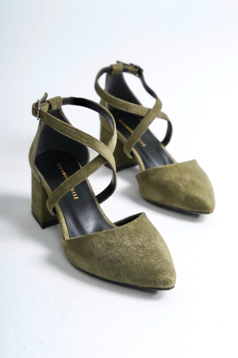 May include: A pair of olive green suede heeled shoes with criss-cross straps and a pointed toe. The shoes have a block heel and a buckle closure.