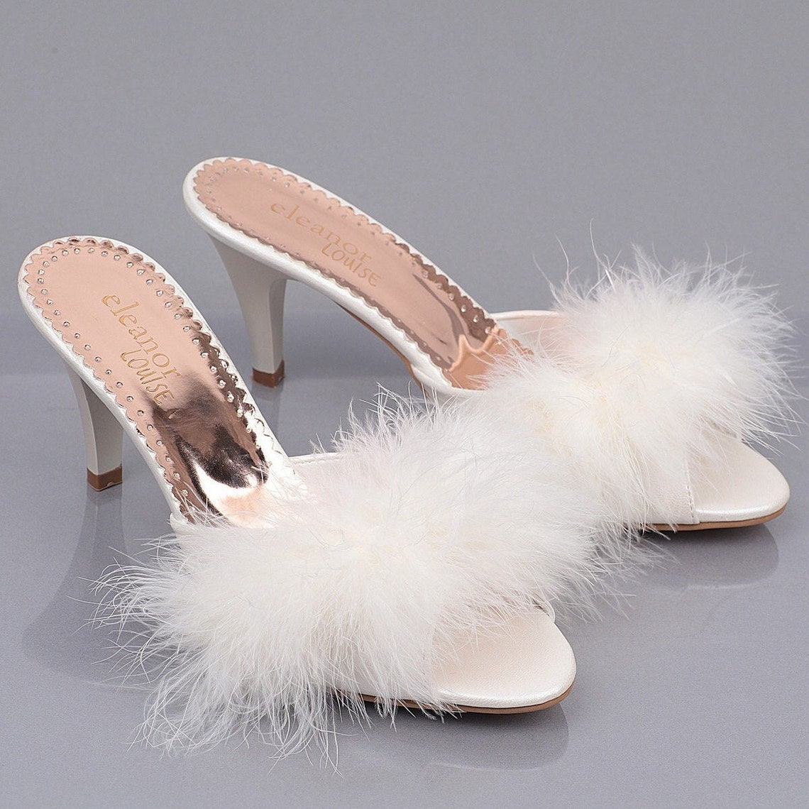May include: Two pairs of white satin heeled slippers with fluffy white feather trim. The slippers have a small gold logo that reads 'eleanor louise'.