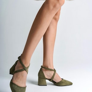 May include: A pair of olive green suede heeled shoes with ankle straps. The straps cross over the top of the foot and buckle at the ankle.