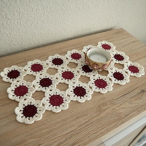 Könnte beinhalten: Handgefertigter gehäkelter Tischläufer mit floralem Design. Der Läufer ist cremefarben mit weinroten Zentren und liegt auf einer Holzoberfläche. Ein kleiner Kerzenhalter mit einer brennenden Kerze befindet sich darauf.
