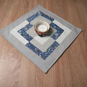 Könnte beinhalten: Ein quadratischer, gesteppter Tischläufer mit grauem Rand und einem zentralen Design aus blauen Blumen- und grau gemusterten Stoffquadraten. Ein kleiner, runder Kerzenhalter mit einer brennenden Kerze befindet sich in der Mitte.