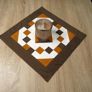 Könnte beinhalten: Ein braun-weiß gesteppter Tischläufer mit geometrischem Muster. Ein klares Glas steht darauf. Der Tischläufer liegt auf einer Holzoberfläche.