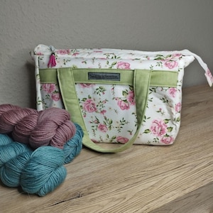 Könnte beinhalten: Eine rechteckige Stofftasche mit weißem Hintergrund und rosa Rosenmuster, akzentuiert durch ein hellgrünes Querband und passende Tragegriffe. Eine kleine rosa Quaste hängt am Reißverschluss. Zwei Wollknäuel liegen daneben.