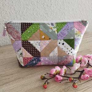 Könnte beinhalten: Ein Patchwork-Reißverschlusstasche in verschiedenen Stoffen und Farben, darunter Rosa, Grün, Braun und Lila. Die Tasche hat einen weißen Reißverschluss und einen Quasten-Zipper. Sie liegt auf einer Holzoberfläche.