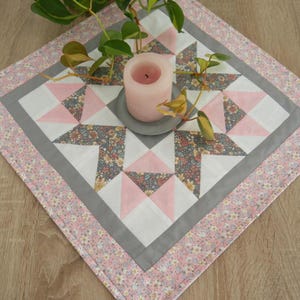 Könnte beinhalten: Handgefertigter, gesteppter Tischläufer mit Sternmuster. Das Design zeigt rosa, weiße und florale Stoffakzente, eingerahmt von einem grauen Rand und einem rosa Blumenrand. Eine rosa Kerze steht darauf.