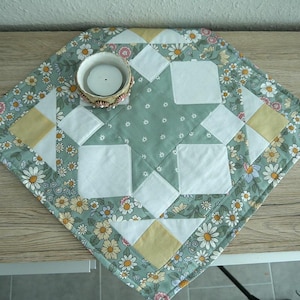 Könnte beinhalten: Quadratischer, gesteppter Tischläufer mit floralem Muster in Grün, Weiß, Gelb und Rosa. Die Mitte zeigt weiße Quadrate und einen kleinen Kerzenhalter mit einer brennenden Kerze.