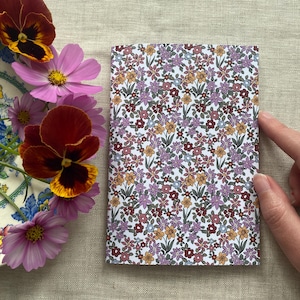 May include: A rectangular notebook with a white cover featuring a dense, repeating floral pattern in shades of purple, yellow, and dark red. Beside the notebook is a decorative plate holding several pink cosmos flowers and dark red pansies.