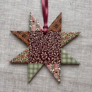 May include: A star-shaped Christmas ornament with a burgundy ribbon. The star is constructed from fabric pieces with diverse patterns, including floral, leaf, and plaid designs. The central square showcases a burgundy floral pattern.