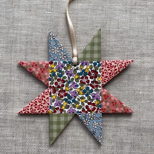 May include: A fabric star decoration with a cream ribbon for hanging. The star is constructed from patterned fabric pieces, featuring floral, gingham, and solid designs. The colour scheme includes red, pink, blue, green, and white.