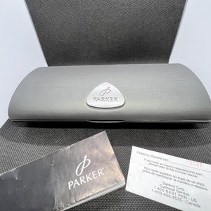 May include: A gray pen case with the Parker logo on the front, along with a black and white pamphlet and a white card with contact information. The case is on a textured surface.