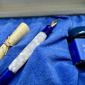 Puede incluir: Una pluma estilogr&aacute;fica azul y blanca con un dise&ntilde;o de m&aacute;rmol blanco y una punta de oro. La pluma est&aacute; en una caja de terciopelo azul con una peque&ntilde;a botella de tinta.