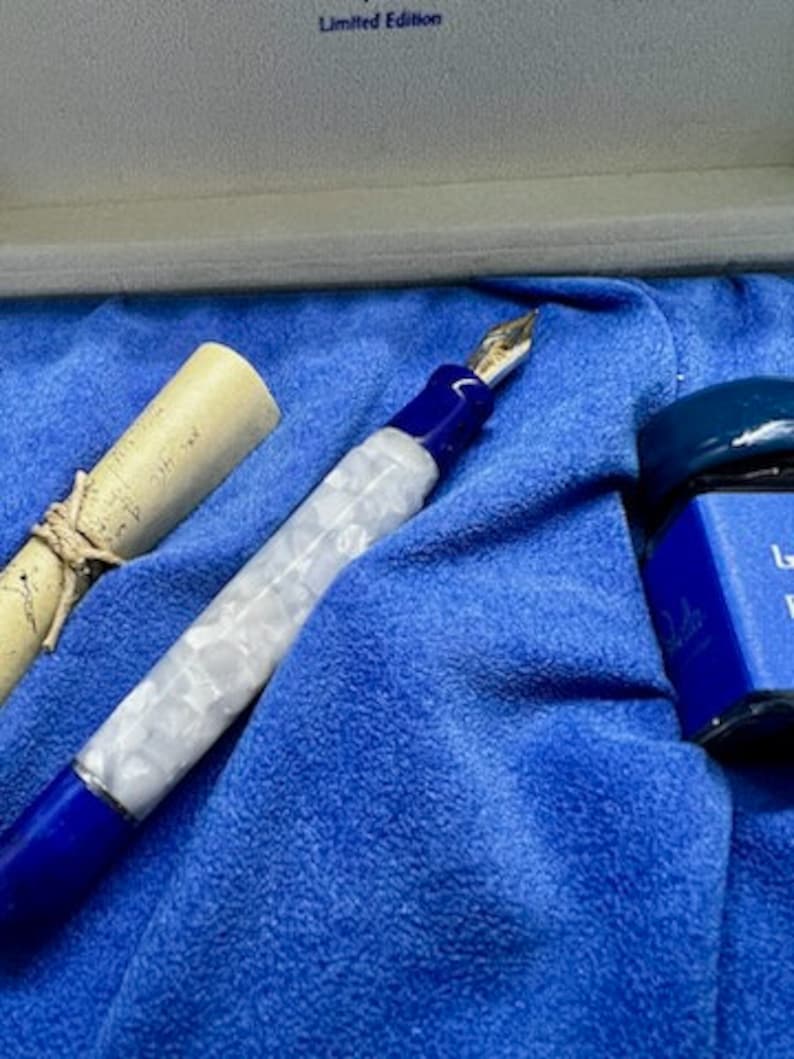 Puede incluir: Una pluma estilogr&aacute;fica azul y blanca con un dise&ntilde;o de m&aacute;rmol blanco y una punta de oro. La pluma est&aacute; en una caja de terciopelo azul con una peque&ntilde;a botella de tinta.