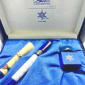 Puede incluir: Una pluma estilogr&aacute;fica azul y blanca con detalles plateados, un rollo enrollado y un frasco de tinta azul con un dise&ntilde;o de estrella de David. La caja est&aacute; forrada de terciopelo azul y tiene el texto "Delta Artigiani della Scrittura Italy" y "Edici&oacute;n limitada" en la tapa.