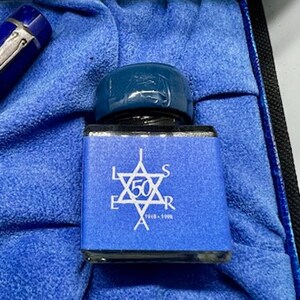 Puede incluir: Un frasco de tinta de vidrio azul con una tapa azul. El frasco tiene una etiqueta blanca con una estrella y el texto "L.E.S. 50 A.R. 1948 - 1998".