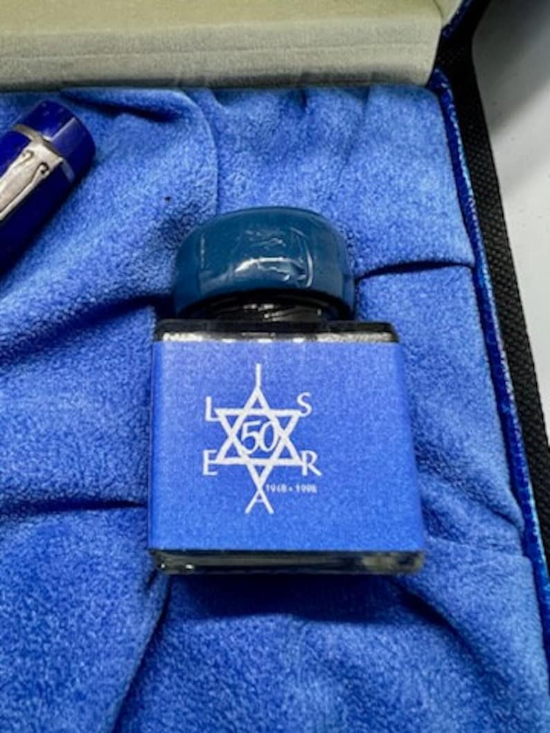 Puede incluir: Un frasco de tinta de vidrio azul con una tapa azul. El frasco tiene una etiqueta blanca con una estrella y el texto "L.E.S. 50 A.R. 1948 - 1998".