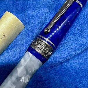 Puede incluir: Una pluma estilogr&aacute;fica azul y blanca con un capuch&oacute;n plateado y un dise&ntilde;o de estrella de David. La pluma tiene la inscripci&oacute;n "CX 50" en el capuch&oacute;n.