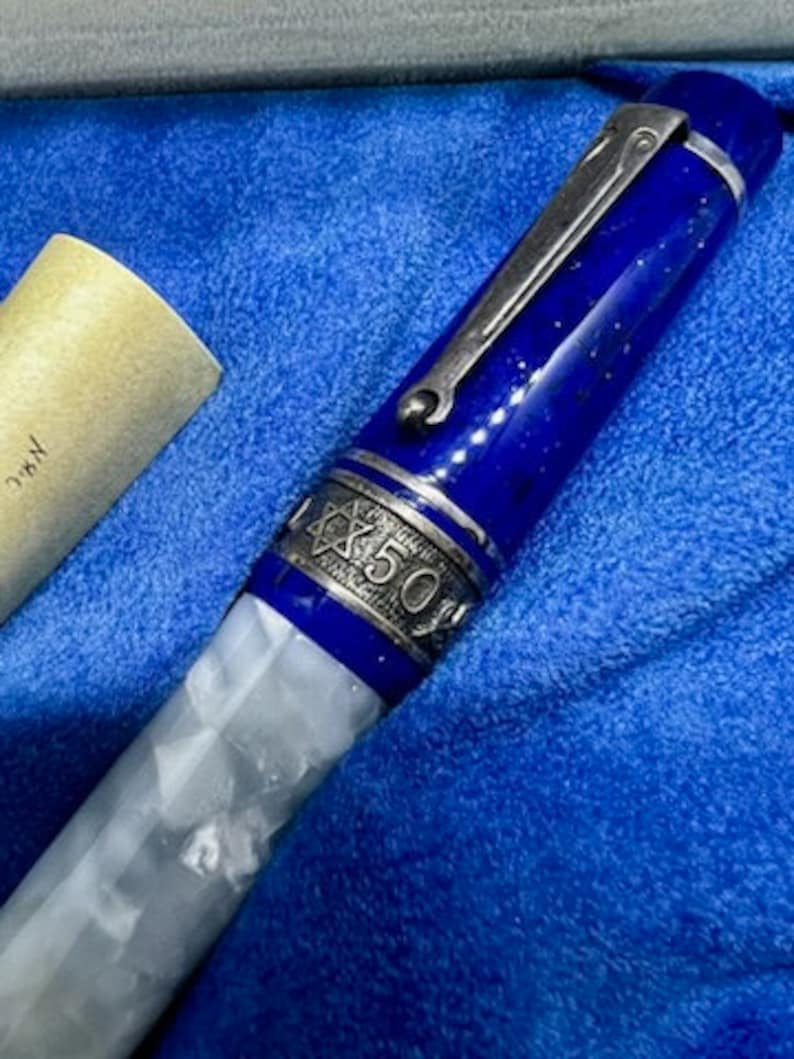 Puede incluir: Una pluma estilogr&aacute;fica azul y blanca con un capuch&oacute;n plateado y un dise&ntilde;o de estrella de David. La pluma tiene la inscripci&oacute;n "CX 50" en el capuch&oacute;n.
