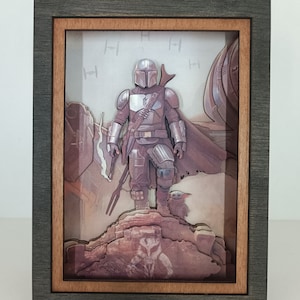 May include: A 3D pop-up art piece in a dark grey frame with a wood-toned inner frame. The art features a Mandalorian character from Star Wars standing on a rock with a small creature beside him. The background is a light grey with a pattern of the symbol for the Mandalorian.