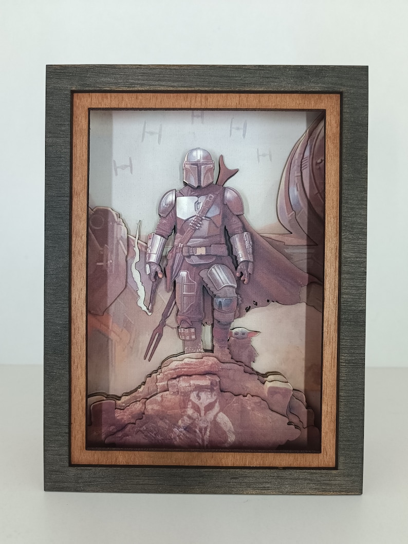 May include: A 3D pop-up art piece in a dark grey frame with a wood-toned inner frame. The art features a Mandalorian character from Star Wars standing on a rock with a small creature beside him. The background is a light grey with a pattern of the symbol for the Mandalorian.