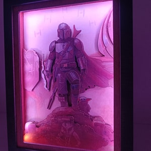 May include: A 3D light-up picture frame featuring a Mandalorian character from the Star Wars series. The character is standing on a rocky surface with a small creature beside him. The frame is illuminated with a pink light.