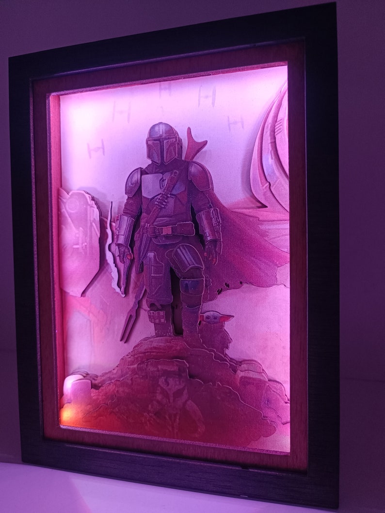 May include: A 3D light-up picture frame featuring a Mandalorian character from the Star Wars series. The character is standing on a rocky surface with a small creature beside him. The frame is illuminated with a pink light.