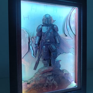 May include: A 3D diorama of a Mandalorian character from the Star Wars universe. The character is standing on a rocky surface with a blue and white background. The diorama is lit from behind, creating a glowing effect.