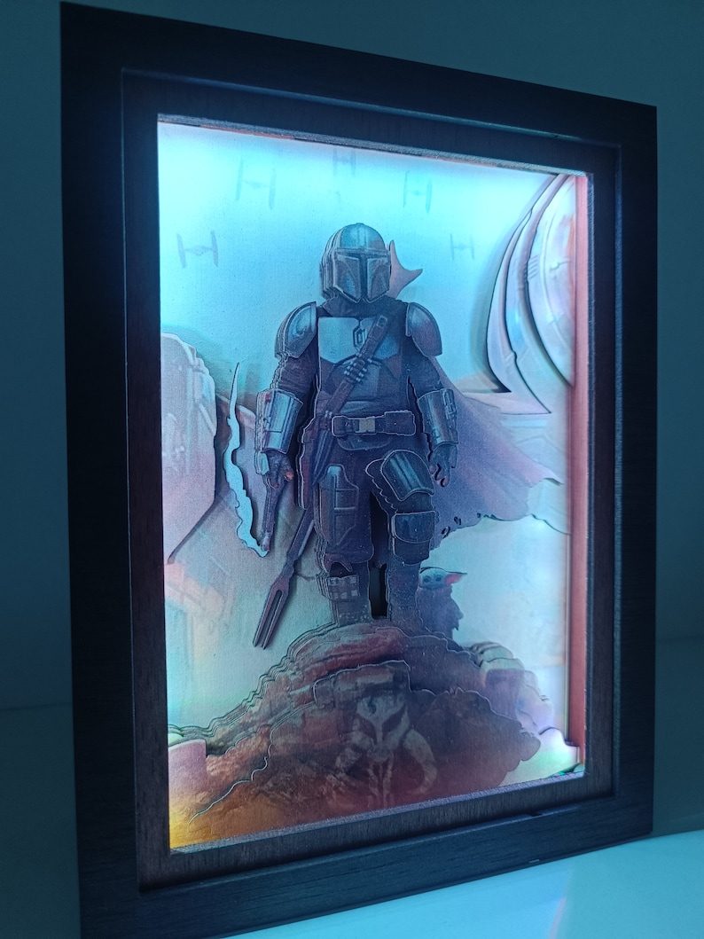 May include: A 3D diorama of a Mandalorian character from the Star Wars universe. The character is standing on a rocky surface with a blue and white background. The diorama is lit from behind, creating a glowing effect.