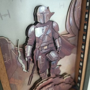 May include: A 3D layered art piece in a wooden frame depicting a Mandalorian character from the Star Wars universe. The character is standing on a rocky surface with a spaceship in the background. The artwork is made of multiple layers of wood and painted in shades of brown, grey, and white.