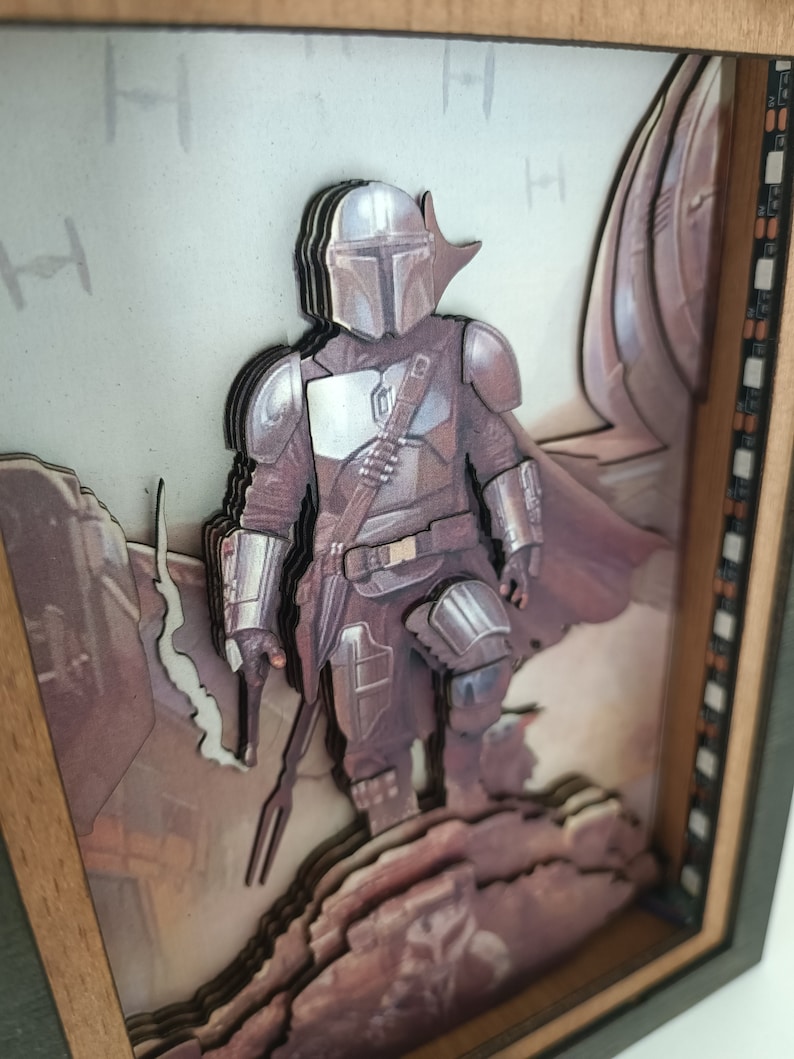 May include: A 3D layered art piece in a wooden frame depicting a Mandalorian character from the Star Wars universe. The character is standing on a rocky surface with a spaceship in the background. The artwork is made of multiple layers of wood and painted in shades of brown, grey, and white.