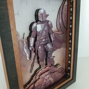 May include: A 3D wooden framed artwork featuring a Mandalorian character from the Star Wars universe. The character is depicted in a layered, multi-dimensional style, standing on a rocky terrain. The background features a blurry image of a spaceship.