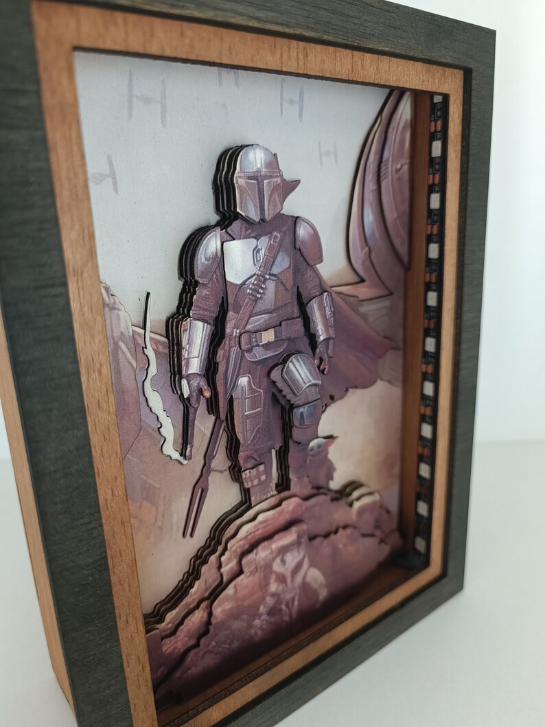 May include: A 3D wooden framed artwork featuring a Mandalorian character from the Star Wars universe. The character is depicted in a layered, multi-dimensional style, standing on a rocky terrain. The background features a blurry image of a spaceship.