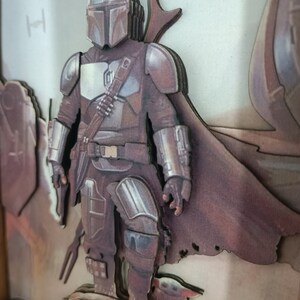 May include: A 3D layered papercraft of a Mandalorian character from the Star Wars universe. The character is standing on a rocky surface with a small creature beside him. The background is a light brown colour.