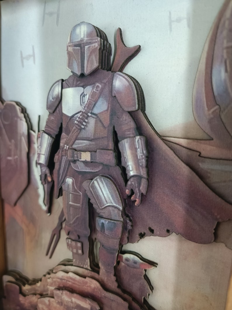May include: A 3D layered papercraft of a Mandalorian character from the Star Wars universe. The character is standing on a rocky surface with a small creature beside him. The background is a light brown colour.