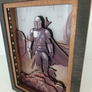 May include: A 3D pop-up art piece in a wooden frame featuring a Mandalorian character from the Star Wars universe. The character is standing on a rocky landscape with a desert background. The frame is dark brown with a light brown inner border.