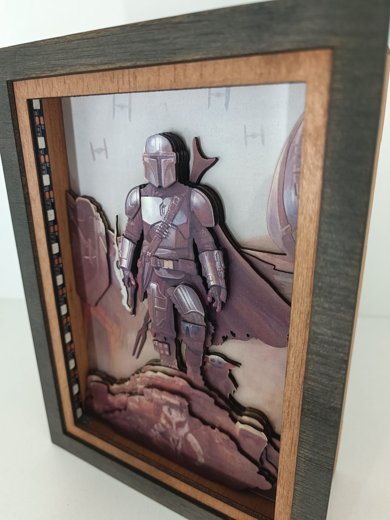 May include: A 3D pop-up art piece in a wooden frame featuring a Mandalorian character from the Star Wars universe. The character is standing on a rocky landscape with a desert background. The frame is dark brown with a light brown inner border.