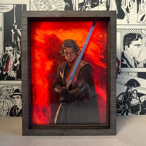 Può includere: Opera d'arte incorniciata raffigurante una figura con una spada laser blu su uno sfondo rosso infuocato. La figura indossa un abito scuro e la cornice è grigio scuro. L'opera è esposta su uno sfondo di fumetti in bianco e nero.