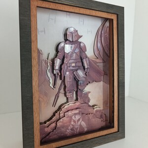 May include: A 3D layered art piece in a dark wood frame with a brown mount. The art features a character from the Star Wars series, The Mandalorian, standing on a rocky cliff with a small creature beside him. The background is a desert scene with a spaceship in the distance.