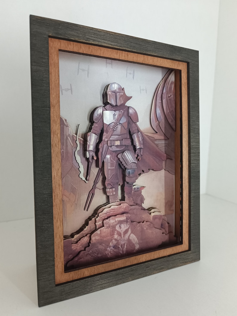 May include: A 3D layered art piece in a dark wood frame with a brown mount. The art features a character from the Star Wars series, The Mandalorian, standing on a rocky cliff with a small creature beside him. The background is a desert scene with a spaceship in the distance.