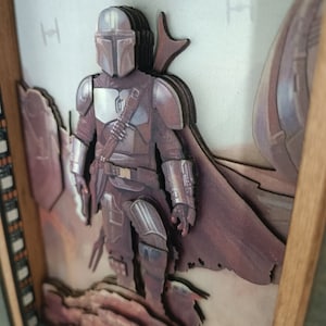 May include: A 3D wooden art piece featuring a Mandalorian character from the Star Wars universe. The character is depicted in a desert setting with a layered, multi-dimensional design.