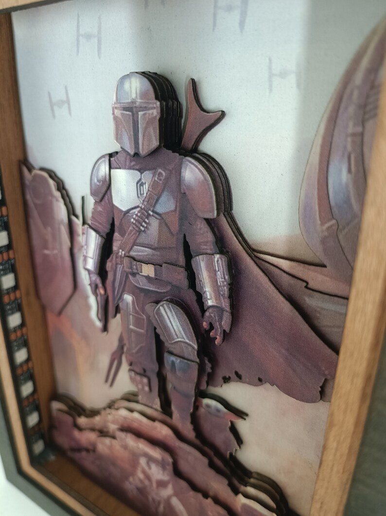 May include: A 3D wooden art piece featuring a Mandalorian character from the Star Wars universe. The character is depicted in a desert setting with a layered, multi-dimensional design.