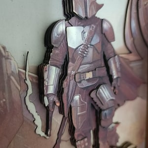 May include: A layered, 3D, wooden art piece of a Mandalorian character from the Star Wars universe. The character is wearing a brown and silver suit with a helmet and a weapon. The background is a brown and beige colour.