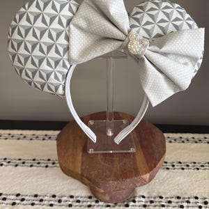 May include: Gray and white geometric pattern mouse ears with a large gray polka dot bow and a rhinestone Mickey Mouse accent. The headband is white, and the ears are displayed on a clear stand.