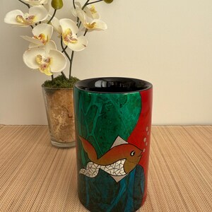 Op de afbeelding: Een cilindrische keramische vaas met een zwarte rand en een kleurrijk ontwerp met een rode, oranje en gouden vis met witte schubben. De vaas heeft een groene, blauwe en rode achtergrond met witte bubbels.