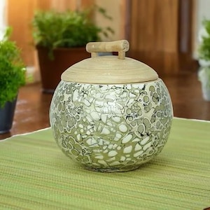 May include: A spherical ceramic jar with a lid, decorated with a speckled pattern in green and white. The lid is a light tan colour and has a small wooden handle. The jar rests on a green placemat, with potted plants visible in the background.