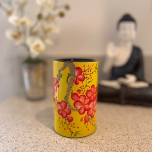 Op de afbeelding: Een gele cilindrische vaas met een bloemenontwerp met roze bloemen en groene bladeren. De vaas heeft een zwarte rand en staat op een wit aanrecht.