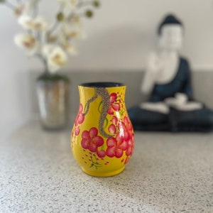 Peut inclure: Un vase jaune avec un motif floral. Le vase a un intérieur noir et est décoré de fleurs rouges et de feuilles vertes. Un accent de paillettes argentées descend sur le côté du vase.