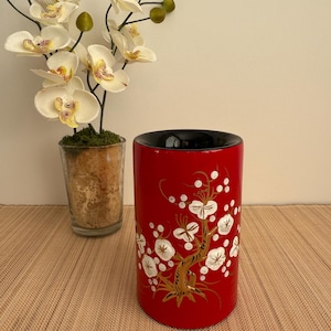 May include: A red cylindrical vase with a black interior, featuring a gold and white floral pattern. The vase is positioned beside a glass container holding a plant and white orchids.